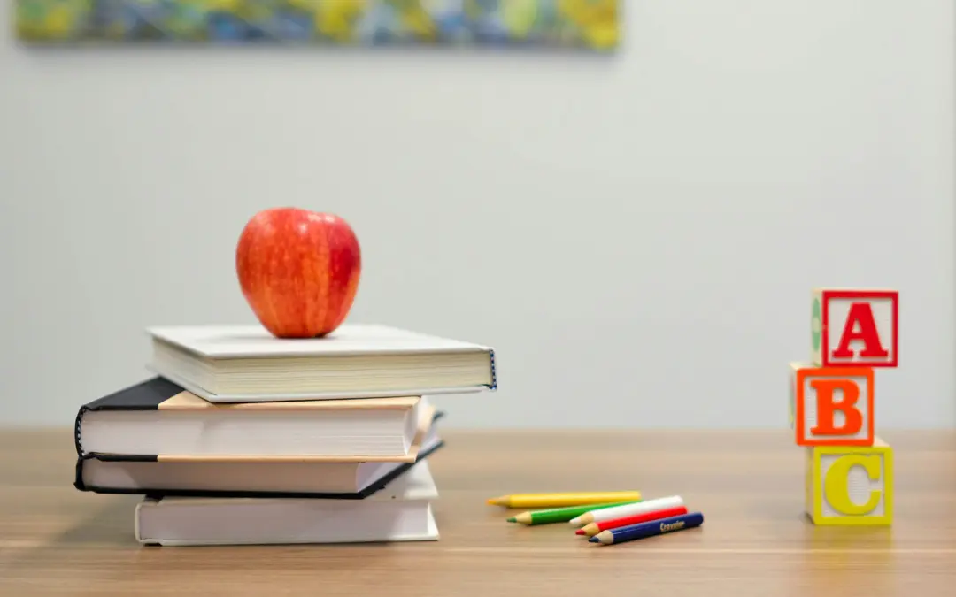 An apple balanced on some books with some children's blocks and crayons next to them.